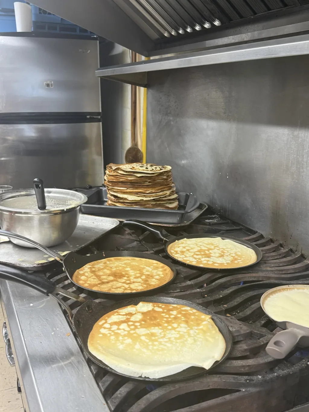 Crepes browning in cast-iron pans on a commercial range, with a finished stack on a tray behind.