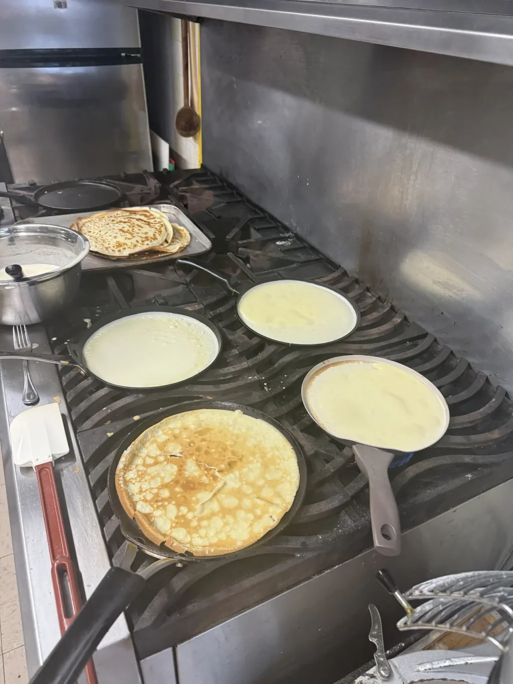 Four crepe pans on a commercial range, batter just poured into three of them.