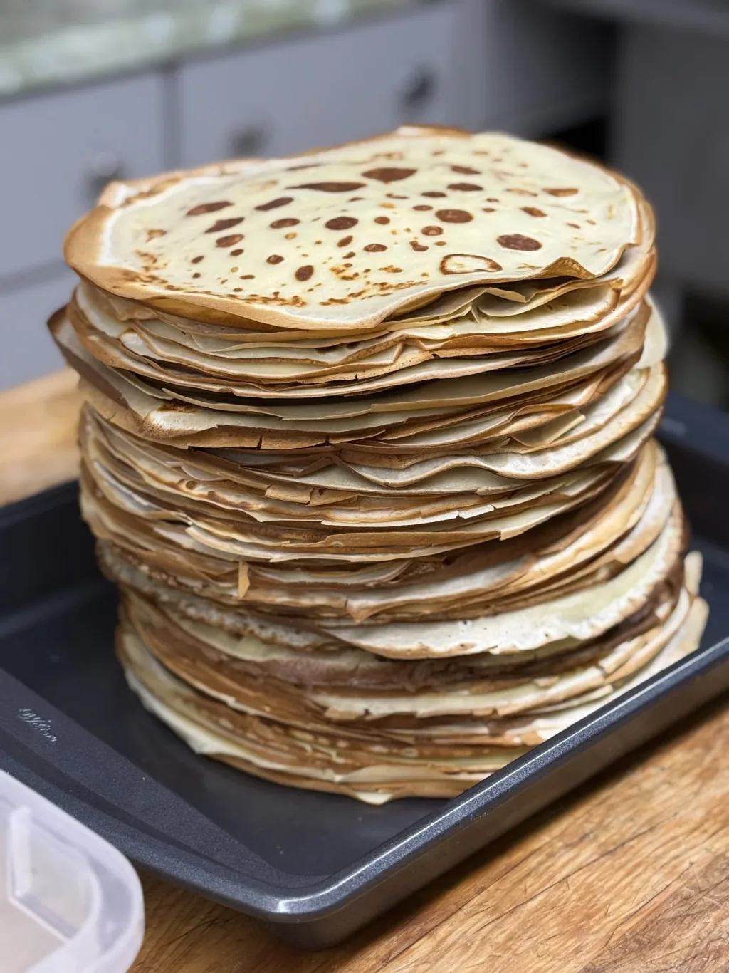 A tall stack of freshly made crepes on a dark baking tray.
