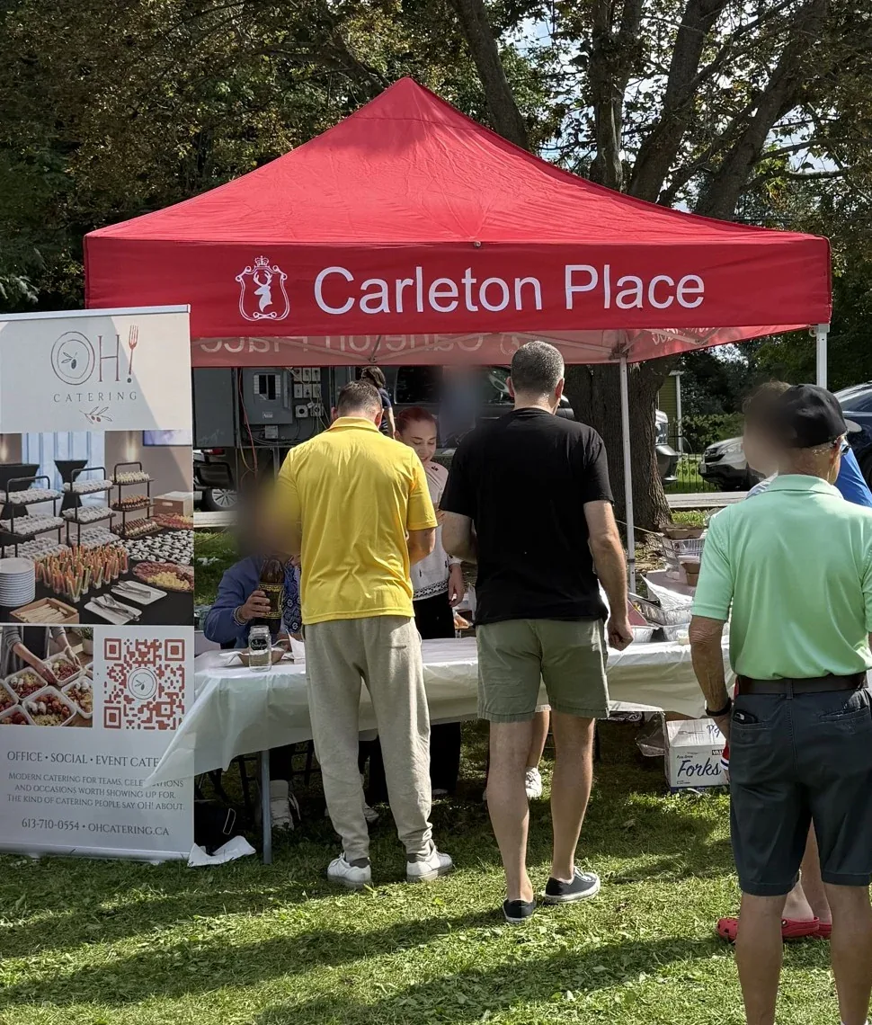 Guests, seen from behind, queue at a red tent beside the OH! Catering banner, in a park.